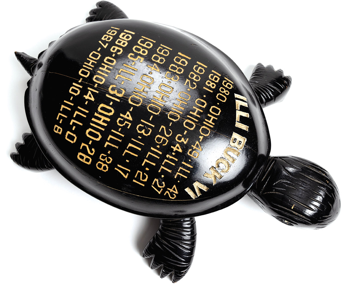 A shiny black turtle carving can be seen from overhead. Its shell lists games the Buckeyes played in the 1980s against Illinois, which were mostly wins. The text says: Illi Buck 6; 1980 Ohio 49, Ill 42; 1981 Ohio 34, Ill 27; 1982 Ohio 26, Ill 21; 1983 Ohio 13, Ill 17; 1984 Ohio 45, Ill 38; 1985 Ill 31, Ohio 28; 1986 Ohio 14, Ill 0; 1987 Ohio 10, Ill