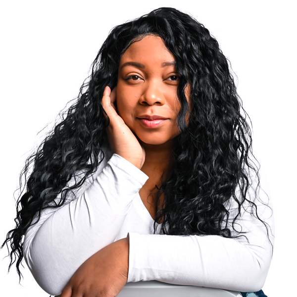 A young black woman with long wavy hair sits in a chair backwards, leaning on its back. She has a close-lipped, confident smile.