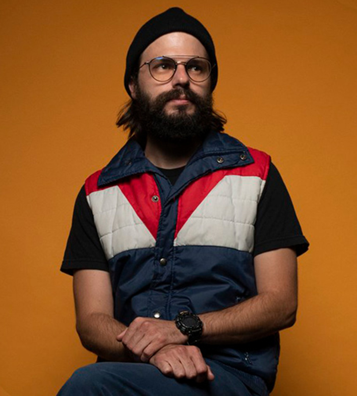 Colin West, a bearded white man with long dark hair, looks contemplatively into the distance. He wears an ’80s-style puffer vest and sits with his hands comfortably crossed on his crossed legs.