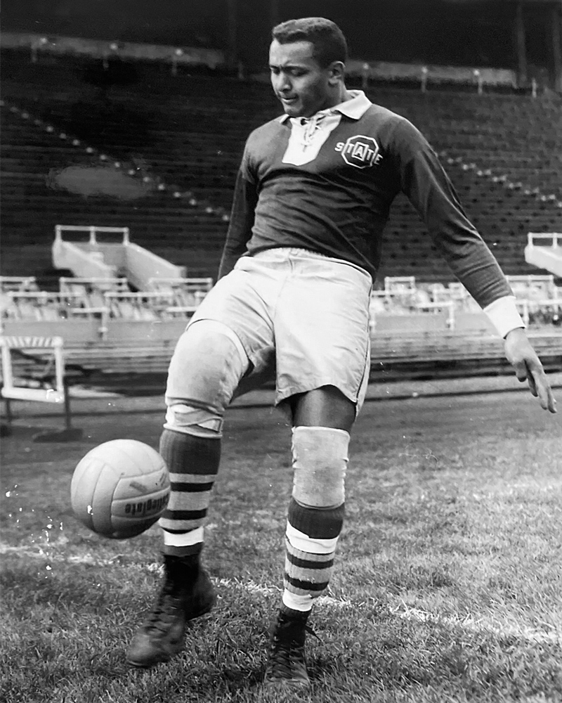 In a historical photo, a young college student with neatly cropped hair juggles a soccer ball. His expression is focused as he stands on one foot, leaning back and raising the other foot in preparation to kick the falling ball back into the air. He wears a long-sleeved uniform shirt, shorts that come close to his knees, knee pads and shin guards under striped socks.