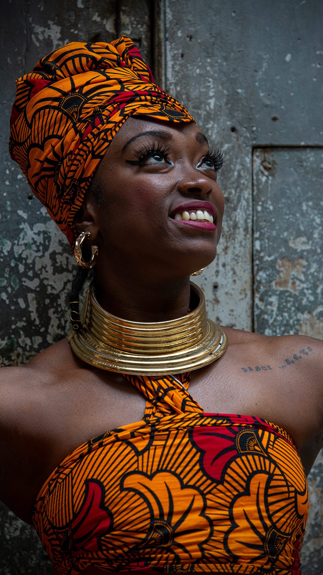 A black woman wearing a matching patterned headpiece and strapless dress looks up happily or hopefully. She has a large, cuff-style gold necklace and a small tattoo across her left shoulder that says Dance … 5, 6, 7, 8. The portrait shows her head and shoulders, not her full body. 