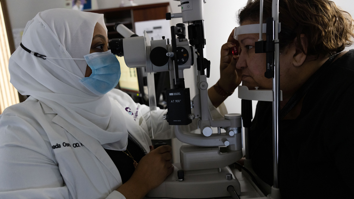 In a darkened room, an eye doctor looks through an instrument into a patient’s right eye while holding a special lens near the patient’s face. The instrument is a slit lamp, a special microscope that helps the doctor examine structures and abnormalities within the eye. The doctor is a black Muslim woman; the patient is a Latina woman.