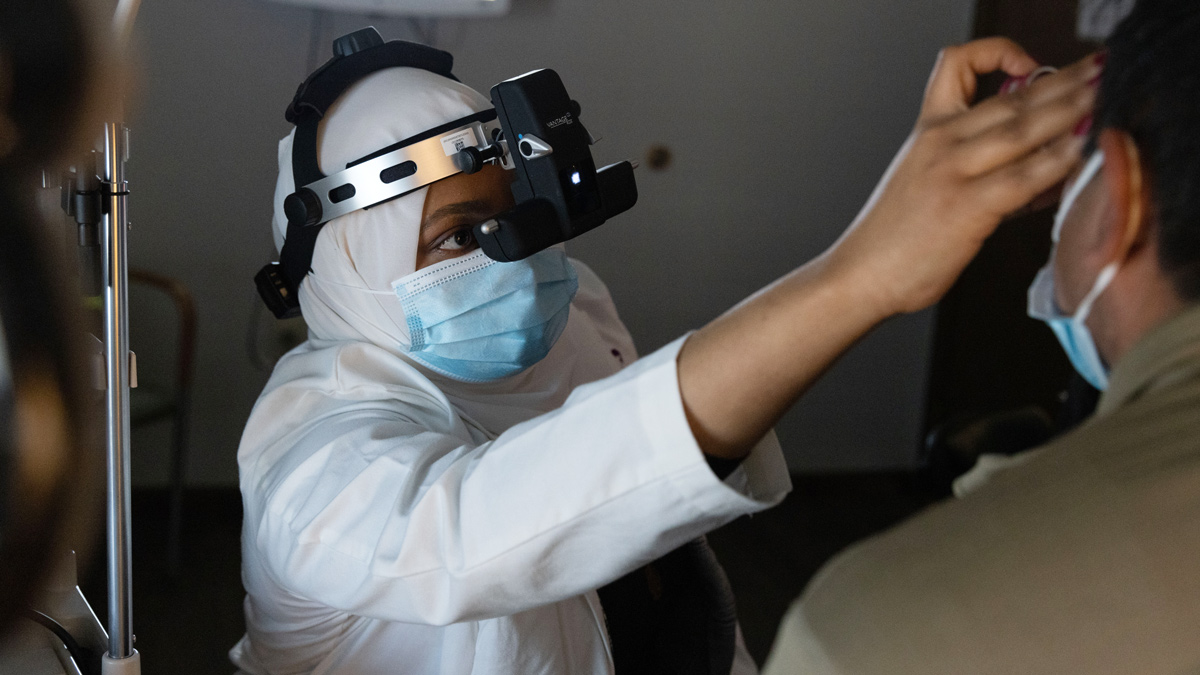 In a darkened room, an eye doctor examines a patient’s left retina by holding a small instrument in front of his eye and wearing a headset that is shiny metal and black plastic. The handheld part has a mirror that reflects light into the patient’s eye; the part on the doctor’s head, called a Binocular Indirect Ophthalmoscope, shows her a closeup view and provides the light.