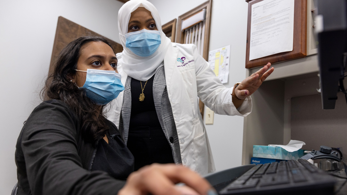 In an office, a doctor gestures as she explains to a student sitting at a computer a point about a patient’s medical records. Both women seem engrossed in their discussion.