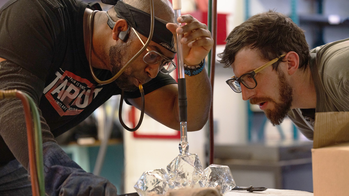 A bearded Black man wearing glasses and a bandana around his head blows into flexible plastic piping connected to a steel blowpipe, which is, in turn, connected to many-sided clear-glass shapes sitting on a worktable. The shapes, representing stars, are connected in a cluster. A white bearded man wearing glasses leans in close to examine the stars. The men’s expressions show a similar determined focus on their work.