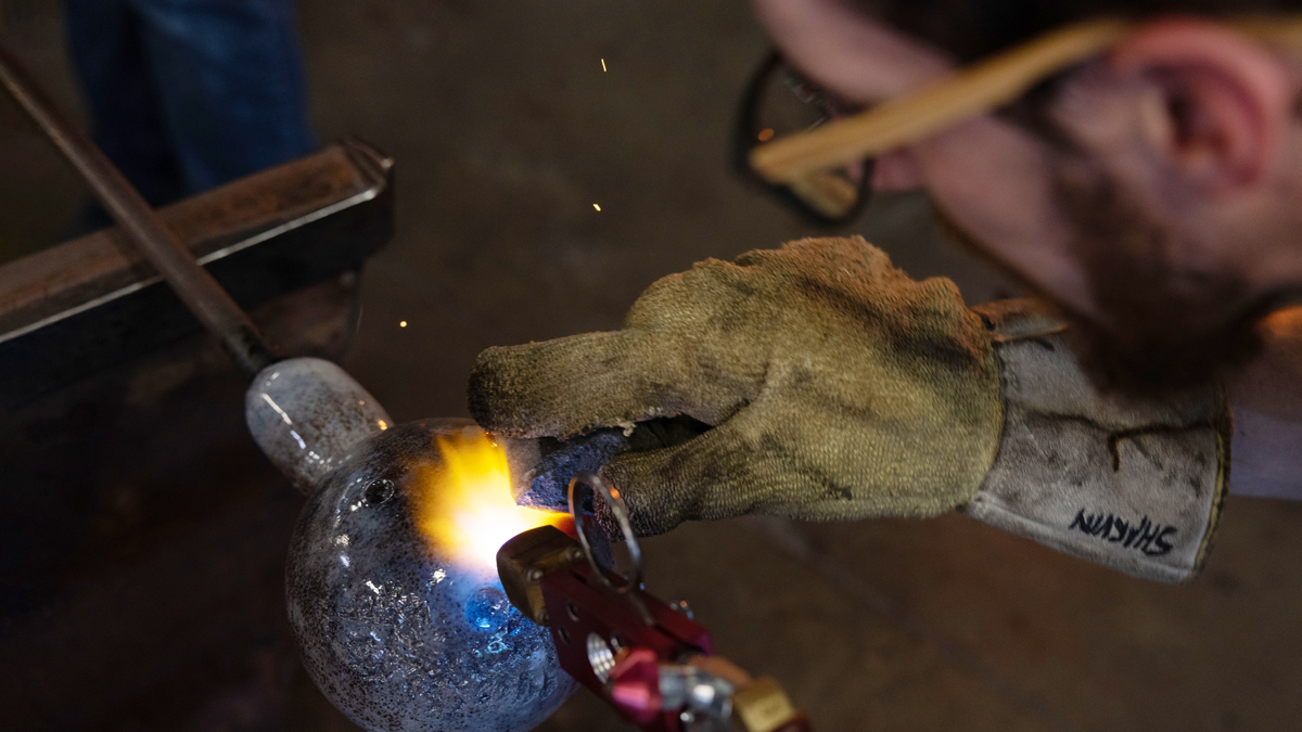 a close-up photo shows, on a worktable, a blowtorch being used to detail a glass ball with blue accents. The person doing the work, who can’t be seen, wears a heat-proof glove to stabilize the hot glass.