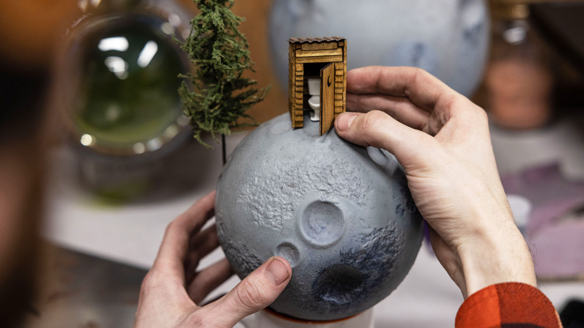 Two hands hold a moon with a tiny outhouse on top, whose door is ajar so a white toilet can be seen inside, and with a pine tree growing off the side. The moon is made of glass, but it is not shiny and smooth. It has craters and is a dull gray. Other moon sculptures are out of focus in the background.