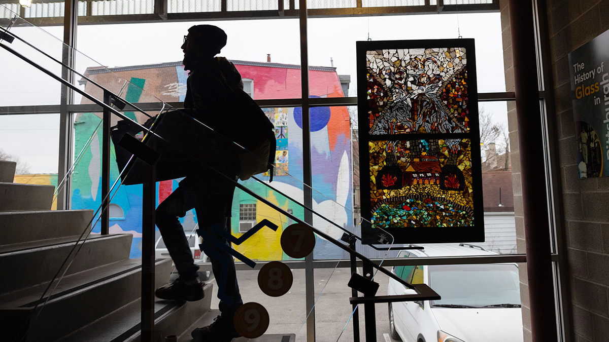 A man climbing stairs and carrying a box of supplies appears as a black silhouette in front of a glass wall. The brightness from outside shines through a couple of elaborately patterned stained glass windows and the building next door is painted with bright colors.