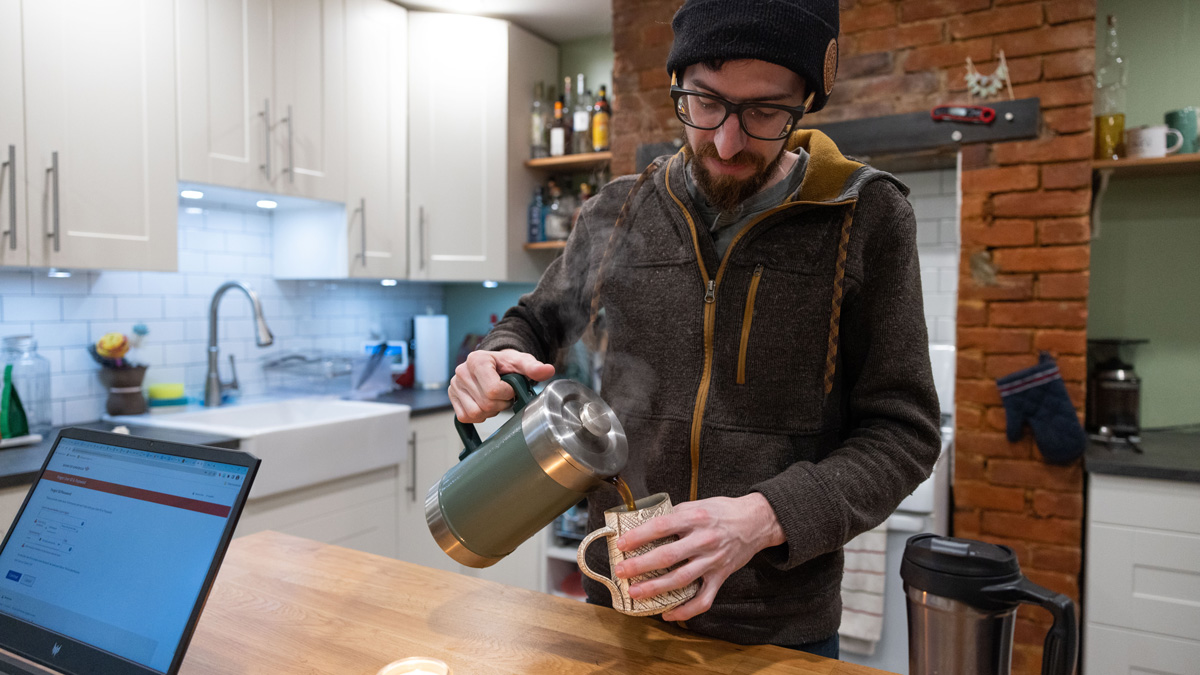 A bearded white man wearing thick-framed glasses, a zip-up and a knitted winter hat pours coffee from a thermos into a hand-made mug. He’s in his kitchen, and in the background, there are a brick wall, white cabinets and shelf with bottles of alcohol.