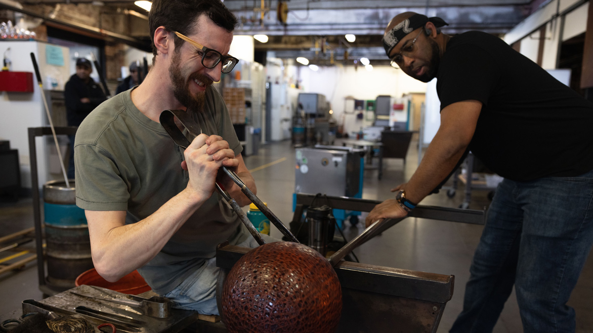 A white man grimaces with effort as he uses giant tongs to stabilize a red-crackled-looking globe with black accents. His work partner, a Black man wearing a bandana, T-shirt and jeans, holds the long pipe attached to the glass globe—it is the tool they used to inflate the globe.