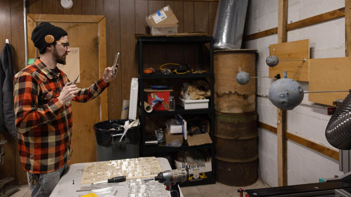 The artist stands in his cluttered personal studio, a small portion of an old auto body shop. He uses his phone to take reference photos of a current project involving grey moon-like glass spheres 