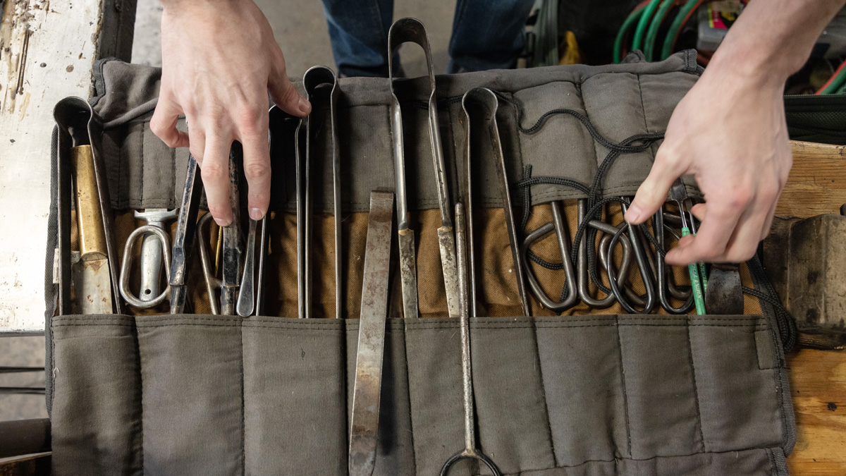 a canvas pouch laid out flat brimming with the metal tools of glass blowing. The craftsmen's hands are visible just above the array.