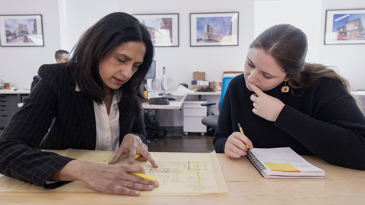 Bhakti works at a table with a member of her company, pointing out details on a large paper she drew an architectural plan on. She and her colleague are both focused on what she’s explaining.