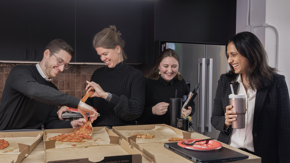 Bhakti and three colleagues stand around a countertop in their office kitchen. Pizza boxes lie open on top, and the colleagues are happily picking out their slices.