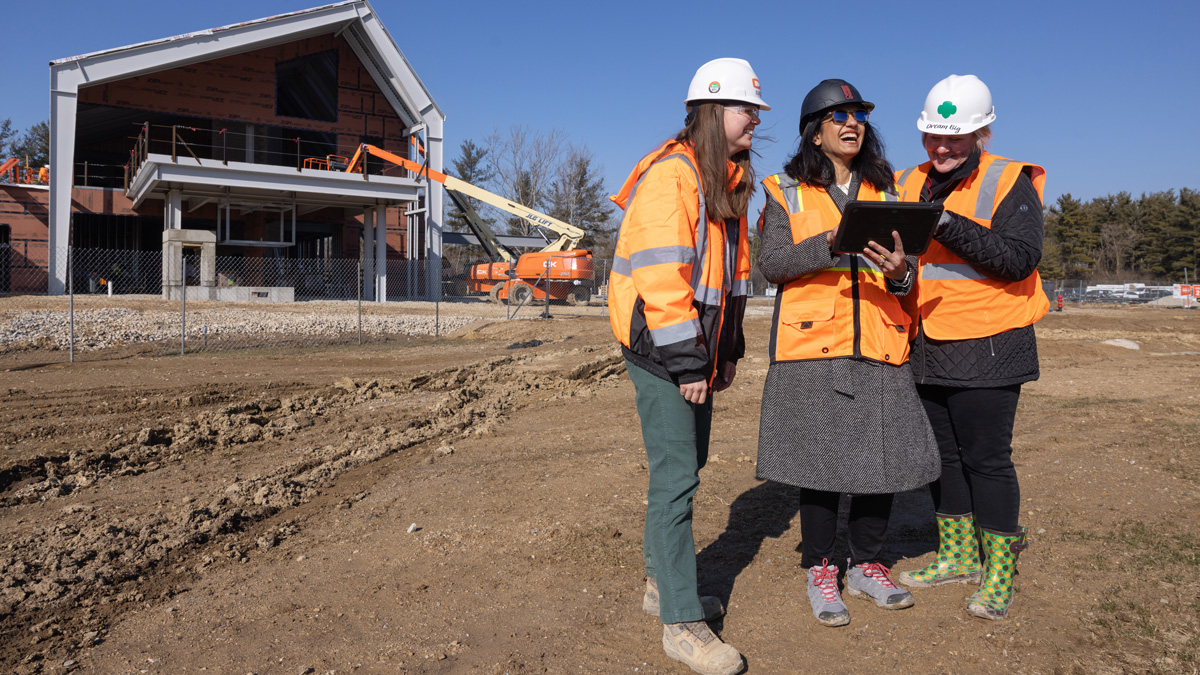 On a sunny day with clear skies, three women stand together looking at a tablet in a large dirt lot. A two story, modern-design building is under construction about 20 yards behind them. All three women wear safety vests and hard hats.