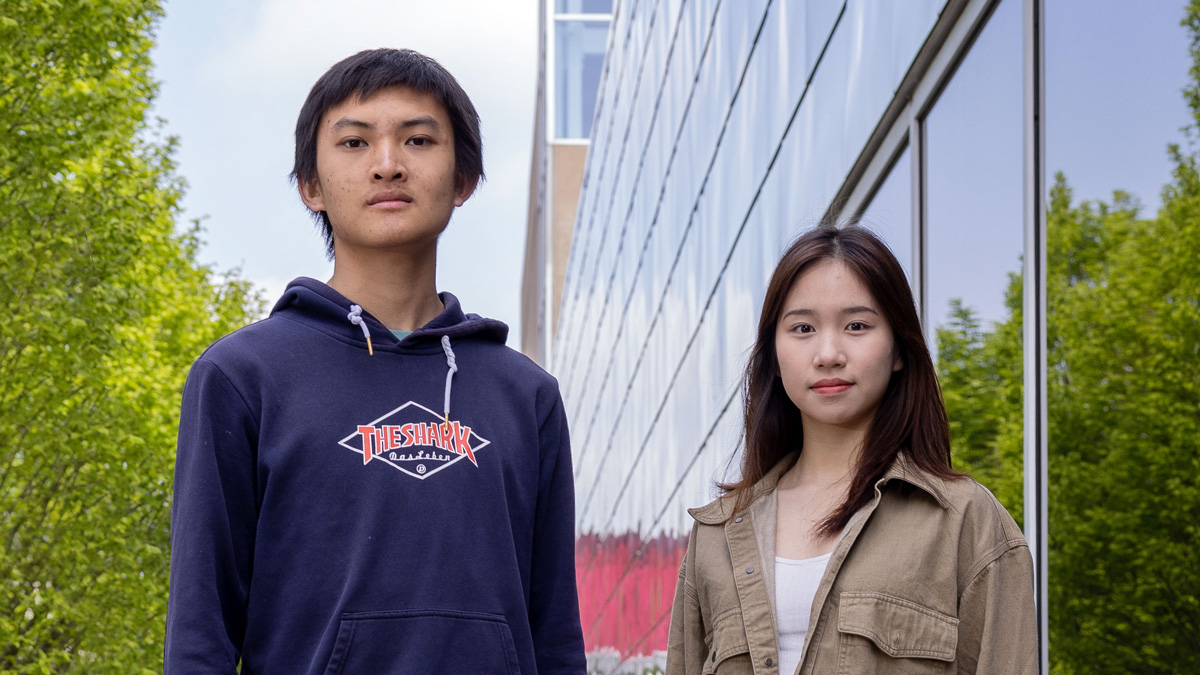 Two college students of Chinese descent pose for a photo next to Ohio State’s recreation building. A young man on the left wears a sweatshirt and sweatpants and a serious look. The student on the right is a young woman with long hair and a slight smile.