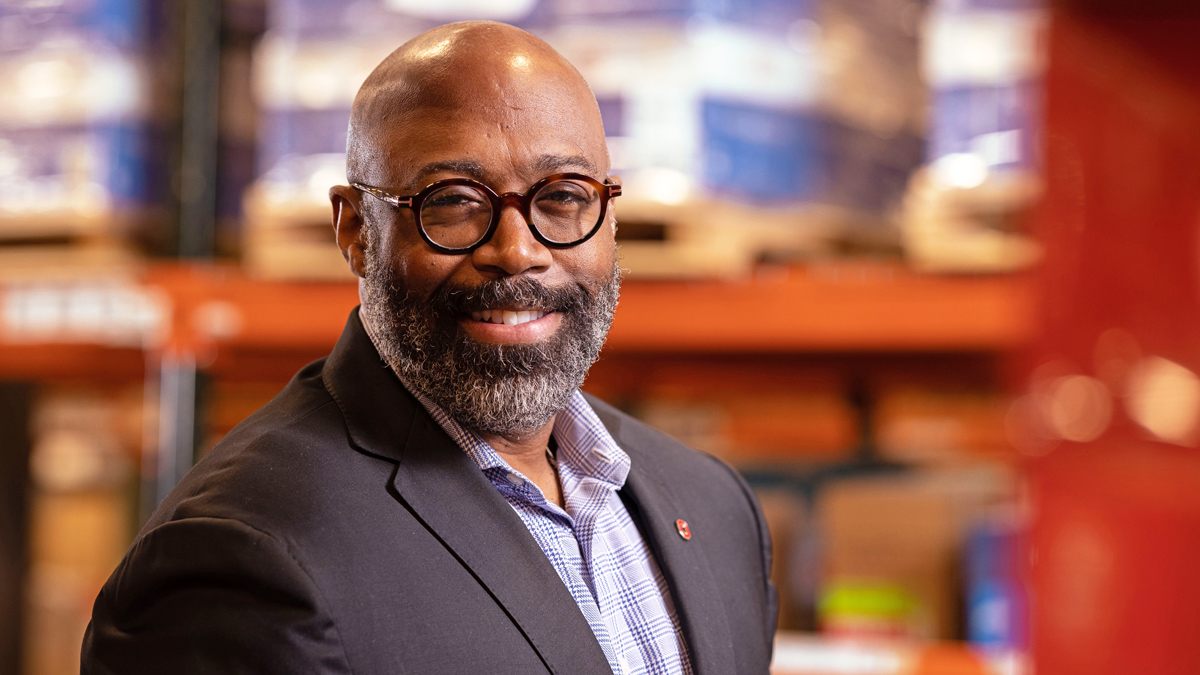 A bearded Black man whose head is shaved wears round glasses and a suitcoat and smiles in a wise way as he stands in a warehouse. His surroundings are out of focus.