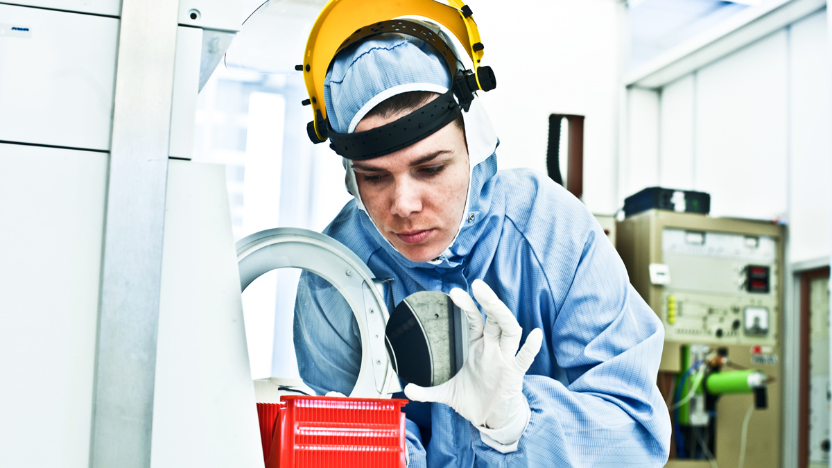 A female worker in a high-tech factory wears a protective suit and leans forward to focus a handheld round mirror on equipment in front of her.