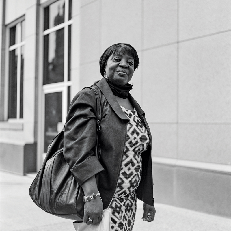 A black woman in a patterned dress and dress coat pauses on a city street. Her hair is sleek, her closed-lip smile is amused.