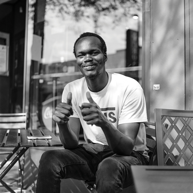 In this black and white photo, a black man gives two thumbs-up and a closed-lip smile as he sits on furniture outside a store or café. He wears a Vans T-shirt.