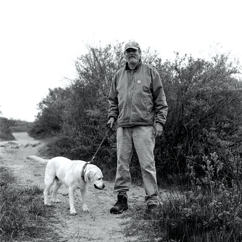 An older white man wears jeans, hiking boots, a Carhart jacket and baseball cap as he walks his yellow Labrador on a sandy path in front of bushes. The setting looks like he might be heading for an ocean, and his expression looks like he’s laughing.
