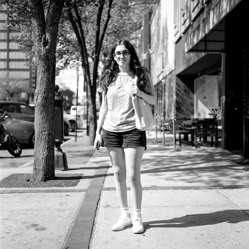 A younger white woman with long dark hair and Harry Potter-style round glasses stands on a city sidewalk in shorts and a T-shirt. Cars are parked diagonally on the street to her right. She offers a closed-lip smile.