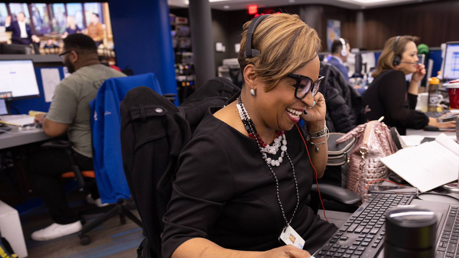 Wearing a headset phone at her work desk, Tracy Townsend chats and laughs. Farther down the line of desks sits Angela An, also wearing a headset phone.