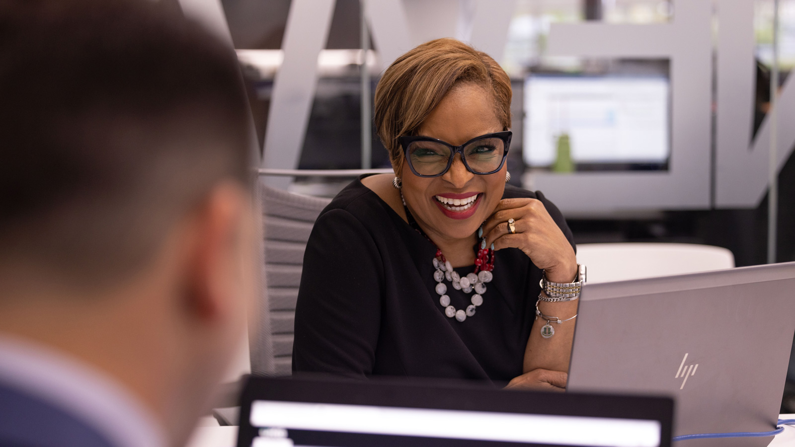 Tracy Townsend looks at a colleague near her at a large table in a meeting room, as she smiles and touches her necklace. Her enjoyment seems both friendly and sincere.