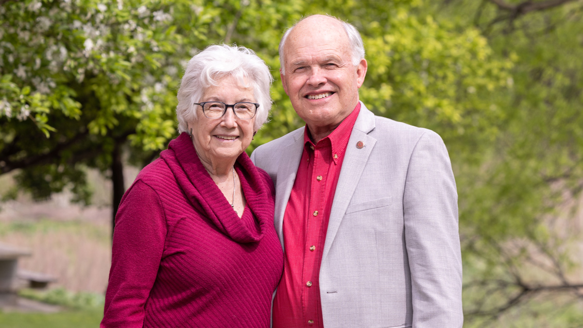 An older man and woman stand close to each other, with their arms wrapped behind each other’s backs. They look friendly and both have white hair, smiles and similar outfits: scarlet shirts and gray pants, though he has added a gray sports jacket. They’re standing outside and trees bloom with spring leaves behind them.