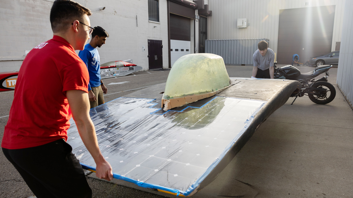 Two men set down the frame of the solar car’s body, while a third stand ready to help. The body is slightly arched with a plastic-looking cockpit. It’s not yet painted or covered with solar panels, although plastic sheeting with a grid covers the part closer to the camera. 