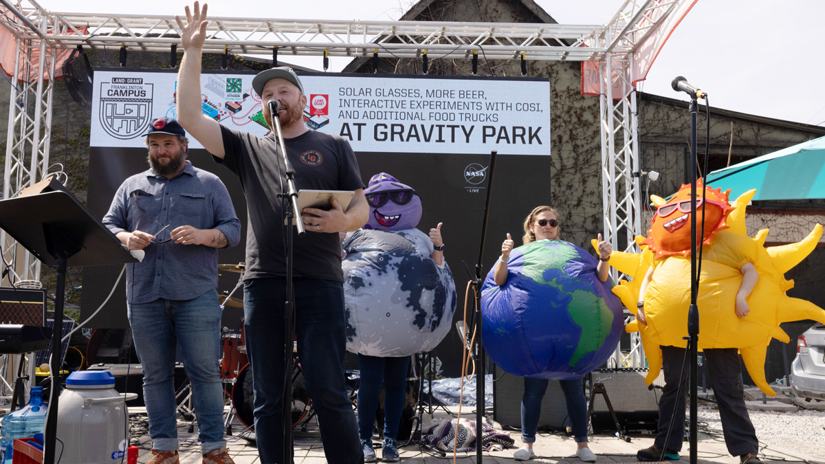 Standing on an outdoor stage, Adam raises one hand as he talks to the crowd. Behind him are Walt and three people dressed up like earth, the sun and the moon.