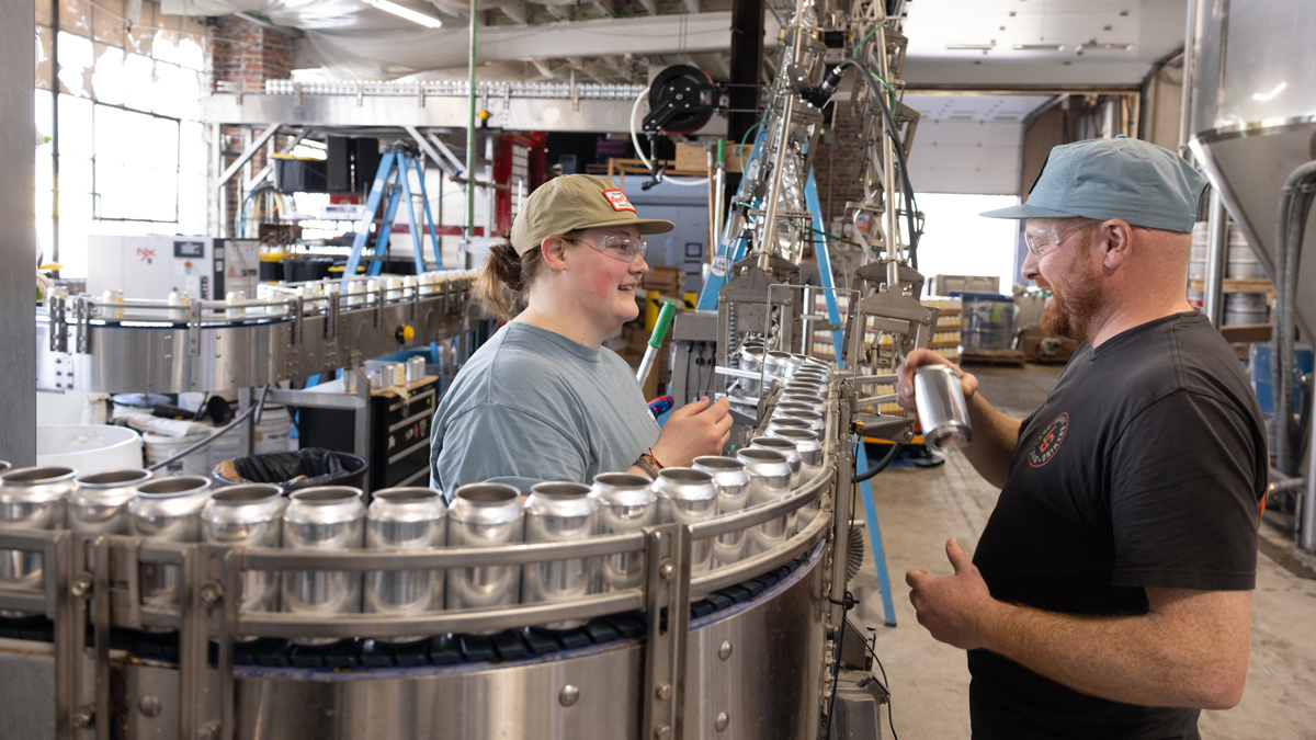 In a factory where a line of shiny but blank cans moves through move on a narrow conveyor belt, a woman wearing a hat and Adam talk while he holds one of the cans. The cans don’t yet have tops and are still empty, presumably as the move along the line to be stamped or wrapped with the brand emblem, filled with beer and sealed closed.