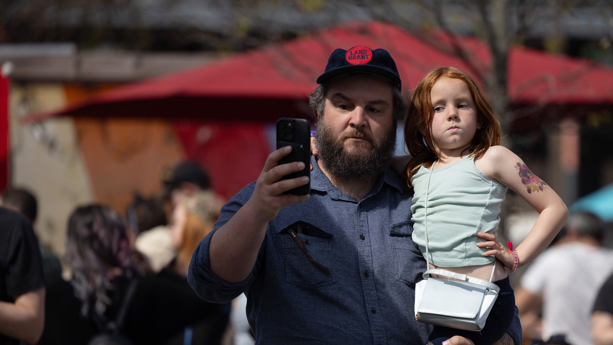 Walt carries his red-headed daughter, who looks to be about 8 years old, as they take a selfie at the eclipse party. He’s wearing a ballcap that says Land-Grant. His daughter puffs out her cheeks, purses her lips, and side eyes her dad’s cellphone camera.