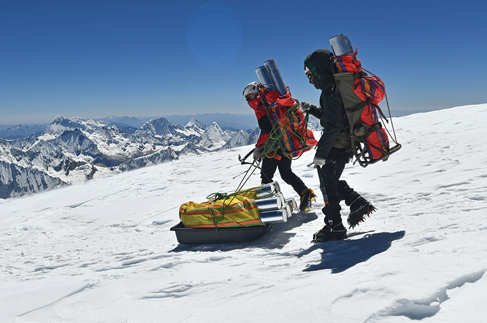 two people pulling a sled that carries multiple ice core samples down the mountain side in the snow