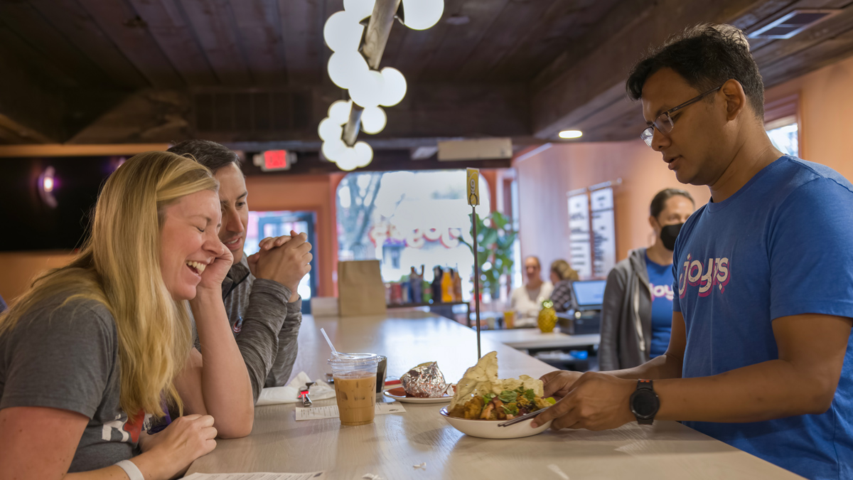 At the counter in a restaurant, a pair of customers laugh as the T-shirt-wearing restaurant owner sets food before them. The closer of the dishes in a bowl piled high.