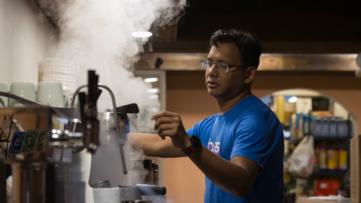 In a dimly lit restaurant in the early morning, steam rises from the espresso machine as a man operates it. He looks a little tired