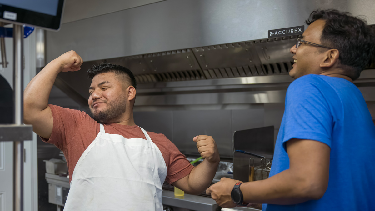 A man wearing a white apron strikes a classic body builder’s pose as another man watche and laughs. Not just his mouth, but his whole body expresses his mirth.