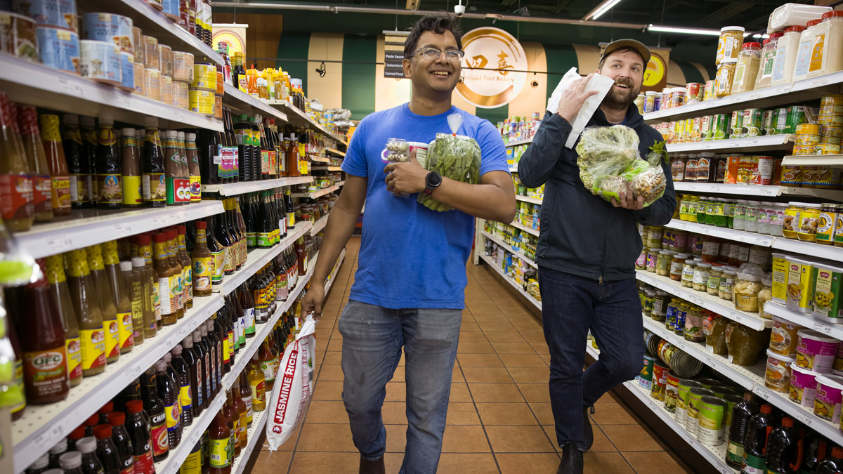 In a grocery store, two men walk up an Asian foods aisle. Both are smiling and their arms are loaded with food, including giant bags of rice and various greens.