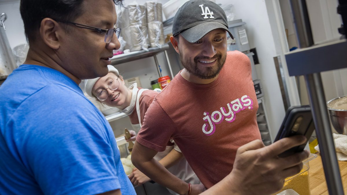 A man shows two co-workers his phone in the kitchen of his restaurant. The co-workers are chuckling.