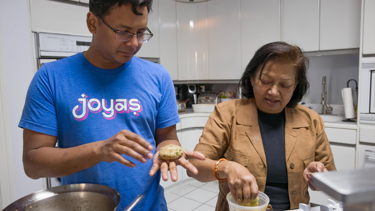 A man of Bangladeshi descent cooks with his mom in her white kitchen. Both are engrossed in the recipe they are testing.