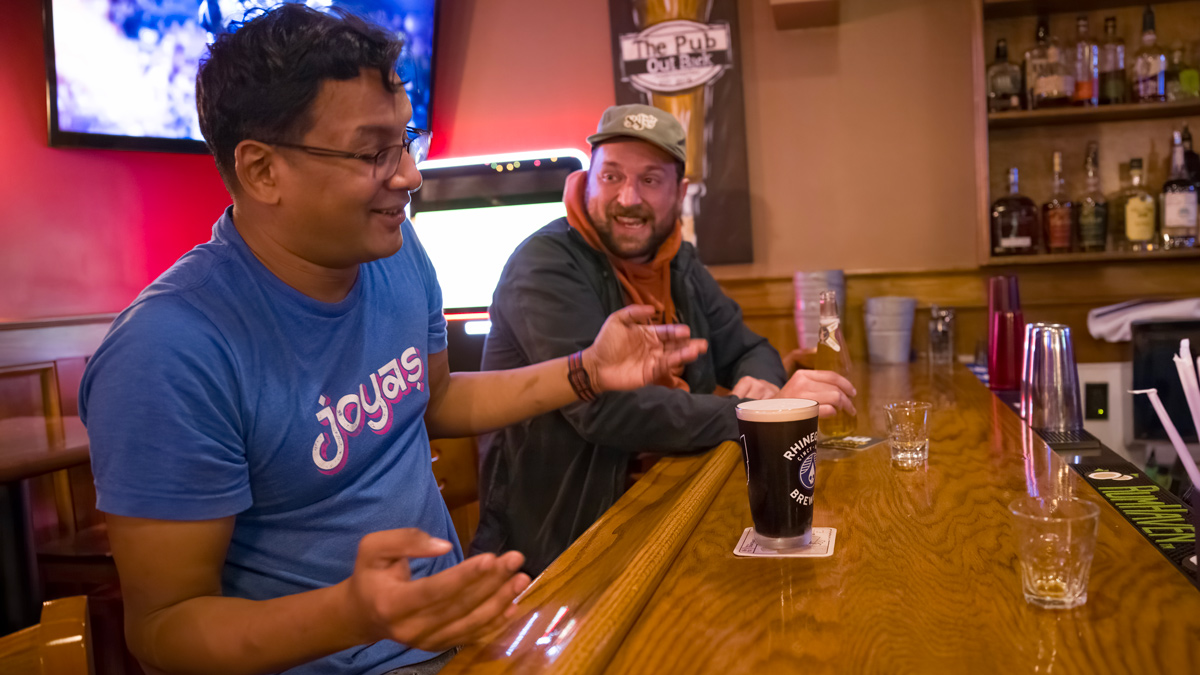 At a bar, a man smiles and shrugs sitting while next to a friend who’s laughing. The first man wears a T-shirt that says “joyas,” the name of his Worthington restaurant. He has a dark beer in front of him and a TV in the background shows a football game.