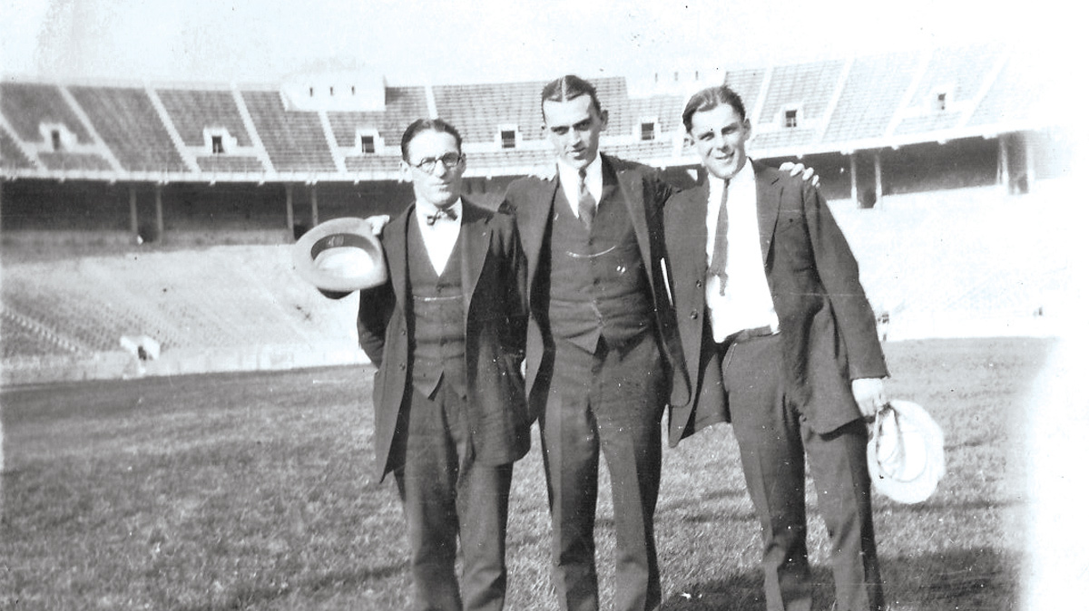 In an old black and white photo, three men in suits with their arms around each other stand on the field inside Ohio Stadium