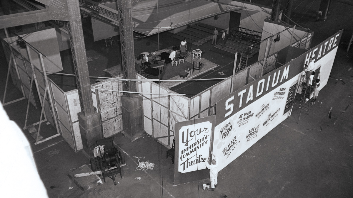 In an old photo taken from overhead, set designers work on a stage setup for a play while others build the theatre in a concourse of the stadium.
