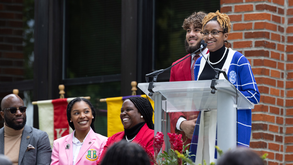 A young black woman wearing a cardigan and glasses smiles as she speaks at the dedication of the NPHC Plaza. Those seated behind her laugh at her remarks.