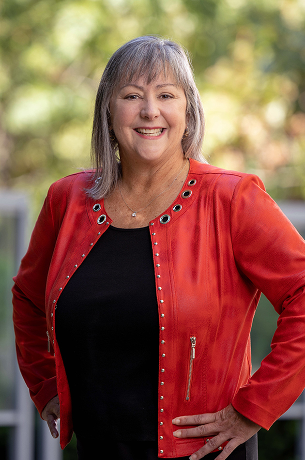 A smiling woman poses outside with hands on hips and a happy smile. Her hair is gray and frames her face; her scarlet jacket is modern and has small studs and zippered pockets.