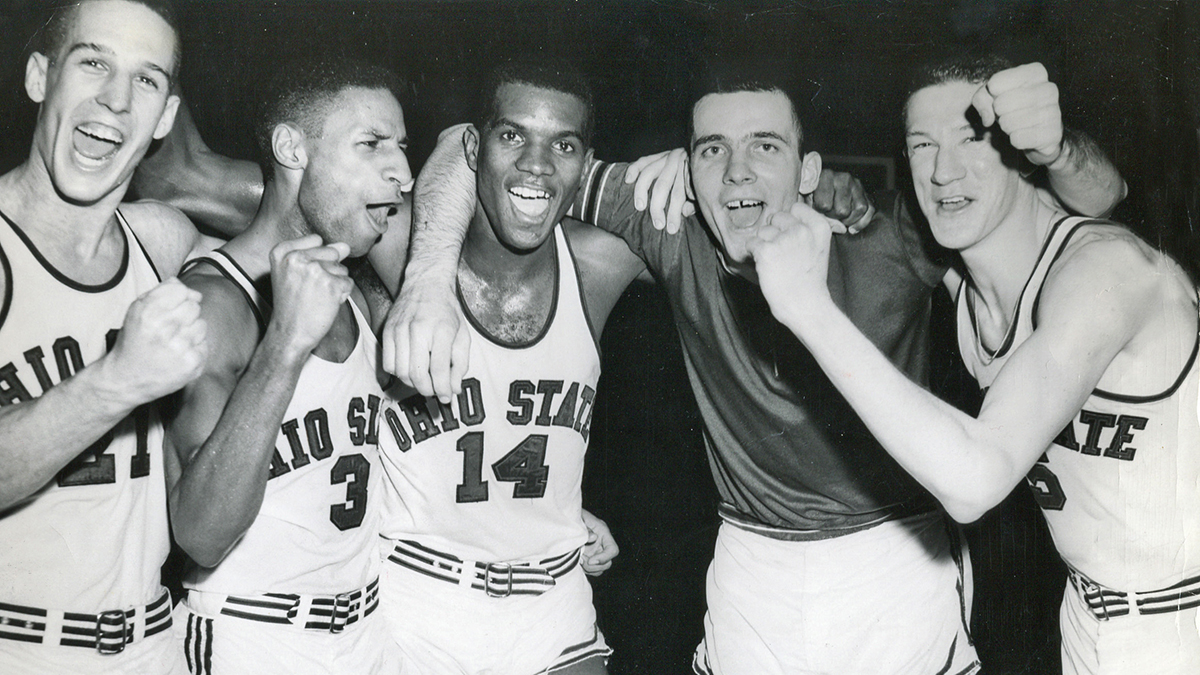 Four baketball players and their coach grin as they pump their fists in celebration after winning a big game. Two of the players and the coach are white and two of the players are black. They all have that clean cut air of the late 1950s and early 1960s.