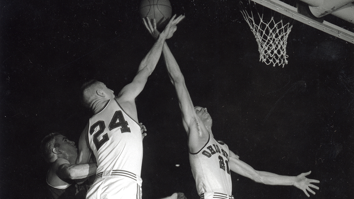 Two Ohio State players, both white and lanky, jump toward the basket. One has a basketball on his fingertips. The other is battling a player from the other team and keeping him from reaching the ball.