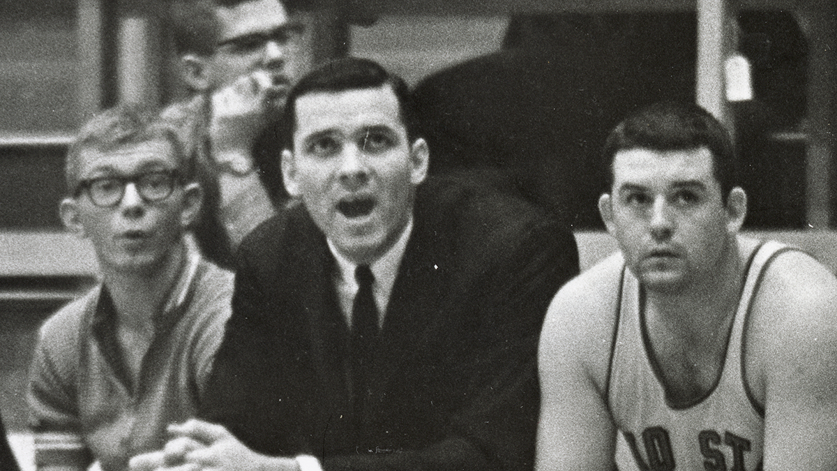 In an old photo, three white men sit on a bench along the basketball court. In the middle is the team’s head coach, a man wearing a suit. To his left is a basketball player. To his right is a young man in a golf shirt. He wears thick glasses and seems to be saying Ooooh!