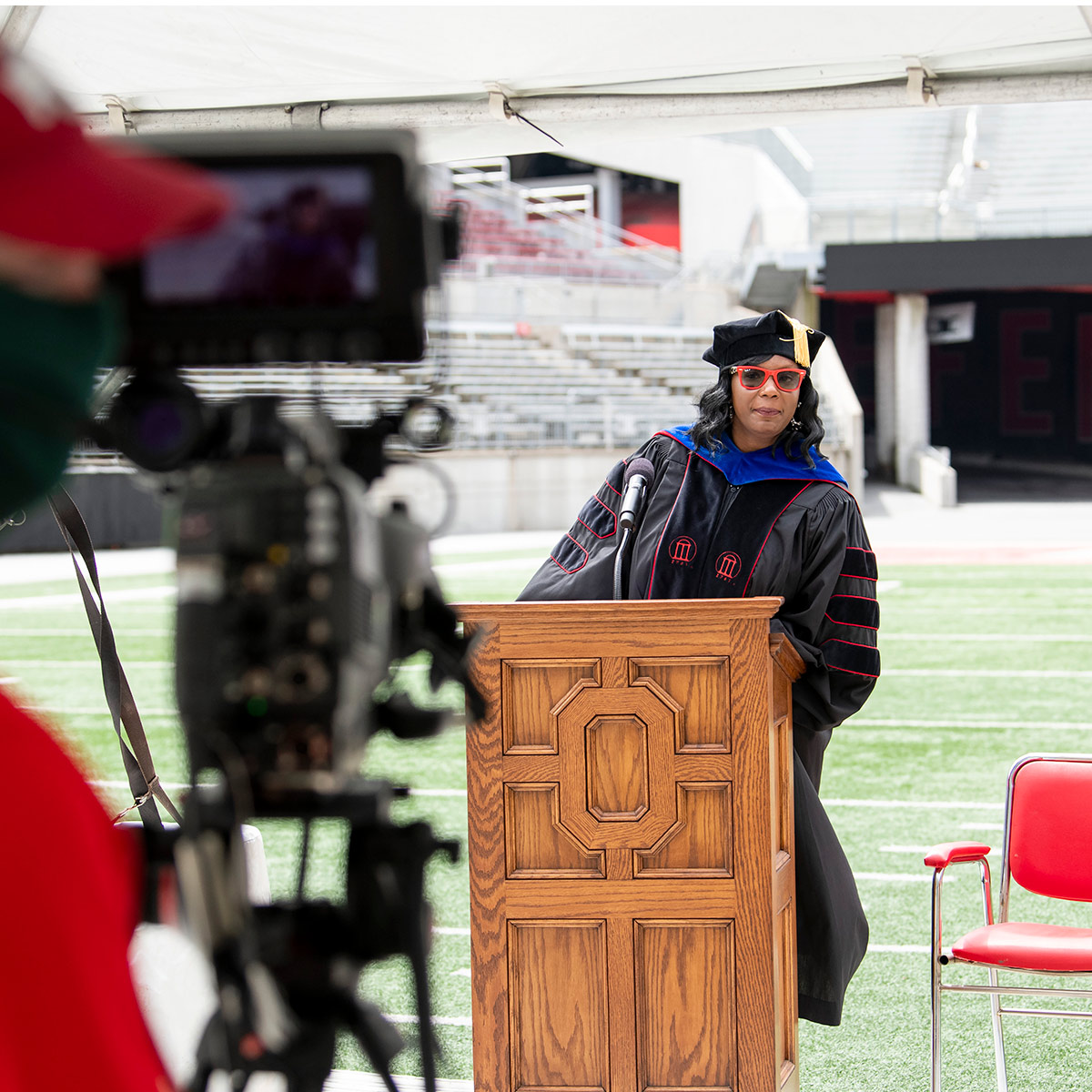 A television camera records a black woman speaking while wearing a cap and gown and red sunglasses.