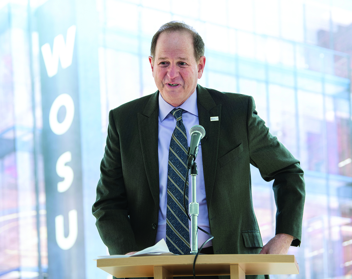 Man in a suit stands in front of a rendering of WOSU studio